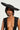 A person exudes vintage glamor in the GIVING EDITORIAL HAT, an oversized wide-brimmed black hat, along with a deep V-neck black outfit against a neutral background. Their ensemble is completed with large hoop earrings featuring cross pendants.