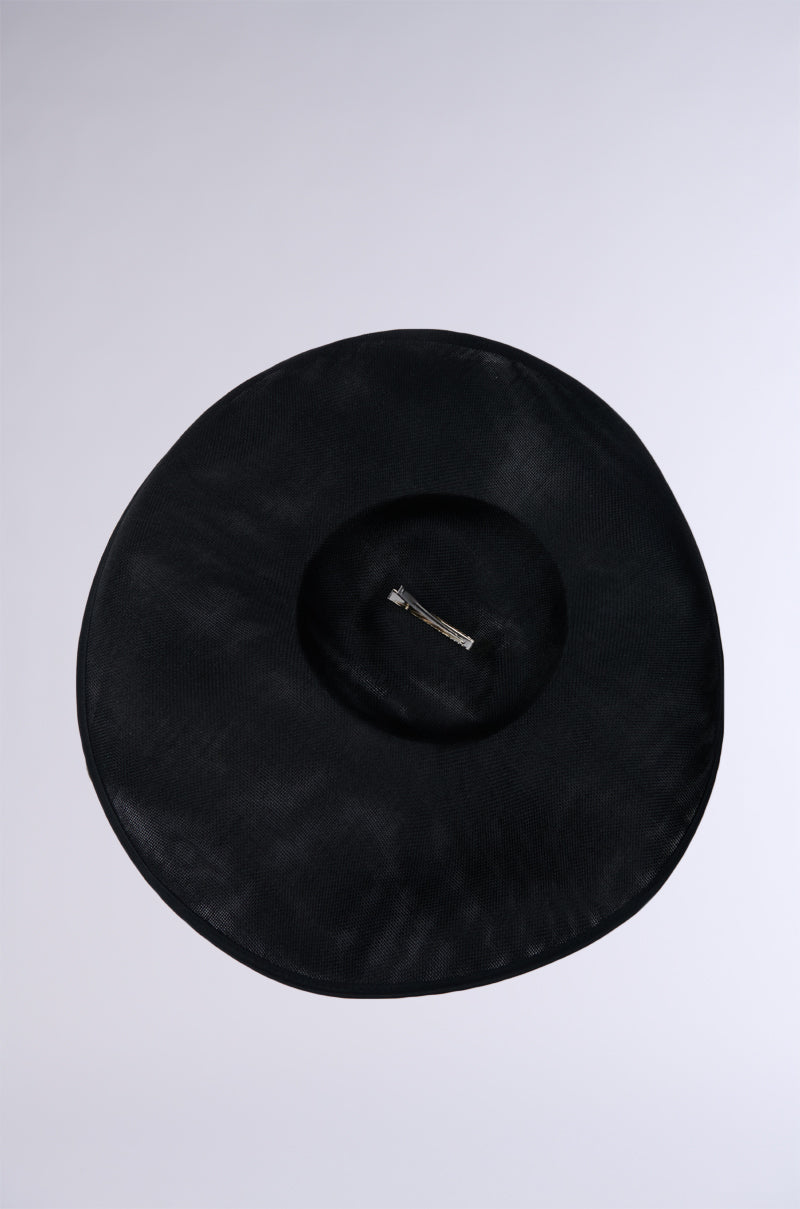 The GIVING EDITORIAL HAT, a round black sun hat featuring a wide brim and radiating vintage glamour, is showcased against a plain, light gray background. A small thin ribbon encircles the inner part of the brim.