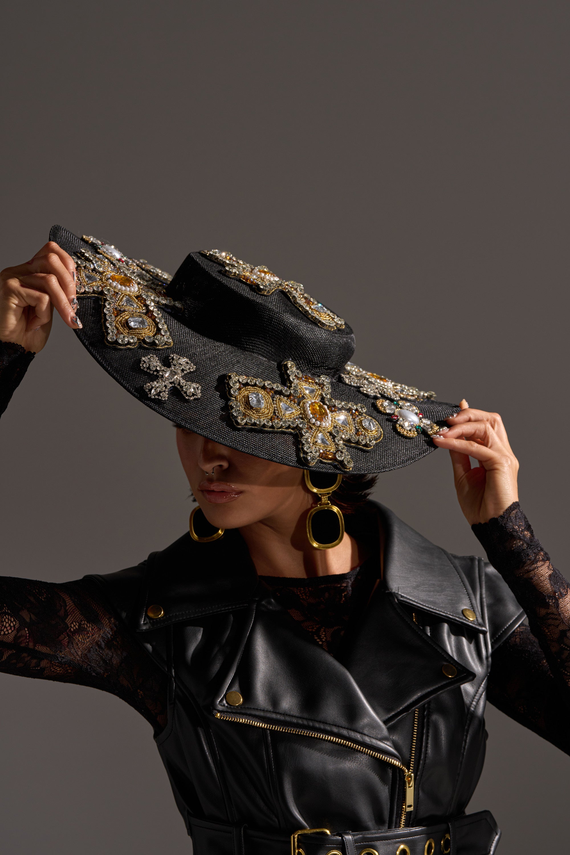 A person in a black lace top and leather vest tilts the STAY BLESSED CLIP ON HAT, which features jeweled embellishments, downward. They wear large gold hoop earrings, set against a gray background.