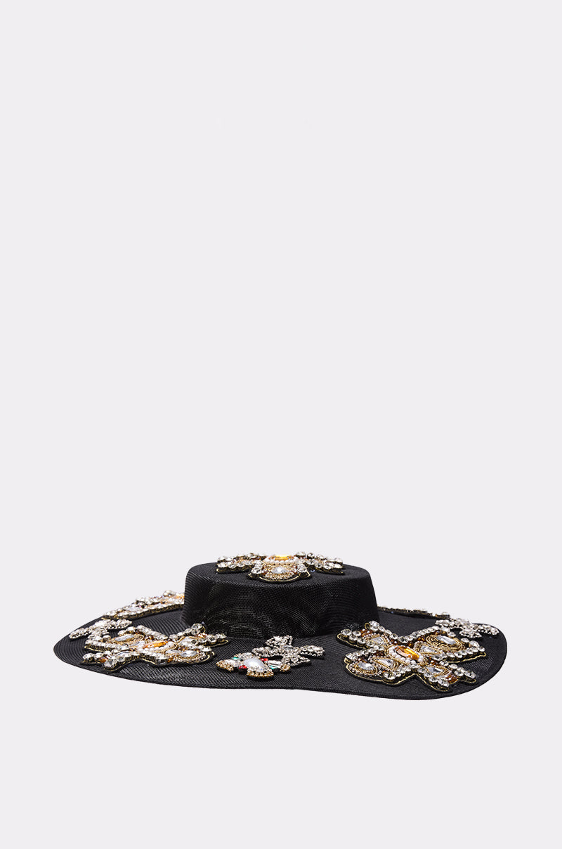 The STAY BLESSED CLIP ON HAT is a wide-brimmed black hat featuring ornate beadwork and jeweled floral embellishments, showcased against a plain light gray background.