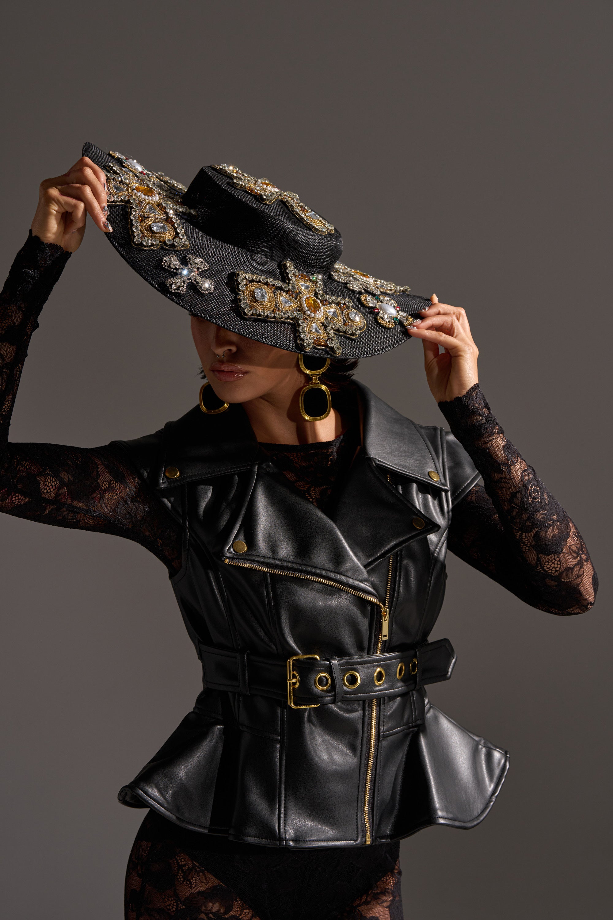 A person in a black lace outfit and belted faux leather vest holds the brim of the STAY BLESSED CLIP ON HAT, a wide-brimmed black hat adorned with jeweled cross appliqués. Large round earrings complete this bold, fashion-forward look.
