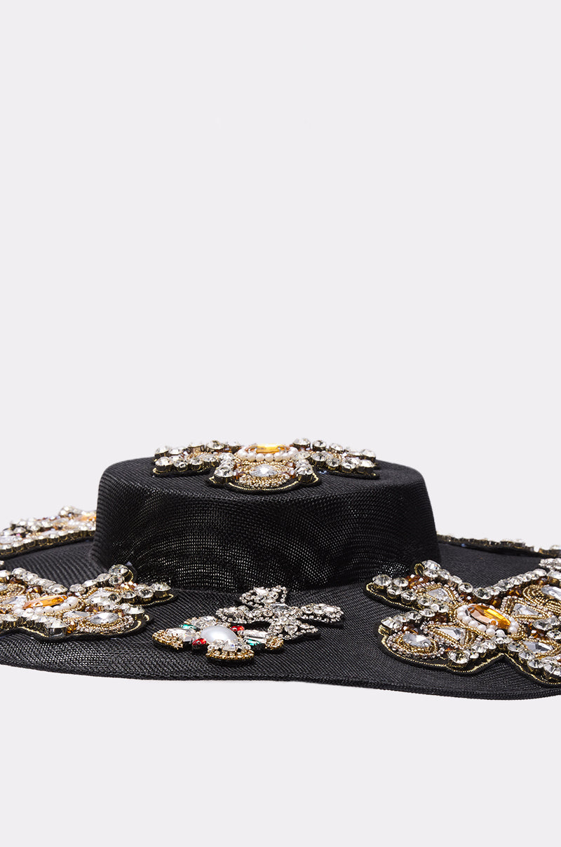 The STAY BLESSED CLIP ON HAT is a black wide-brimmed hat adorned with gold, pearl, and crystal jeweled embellishments, making a bold statement accessory against a plain white background.