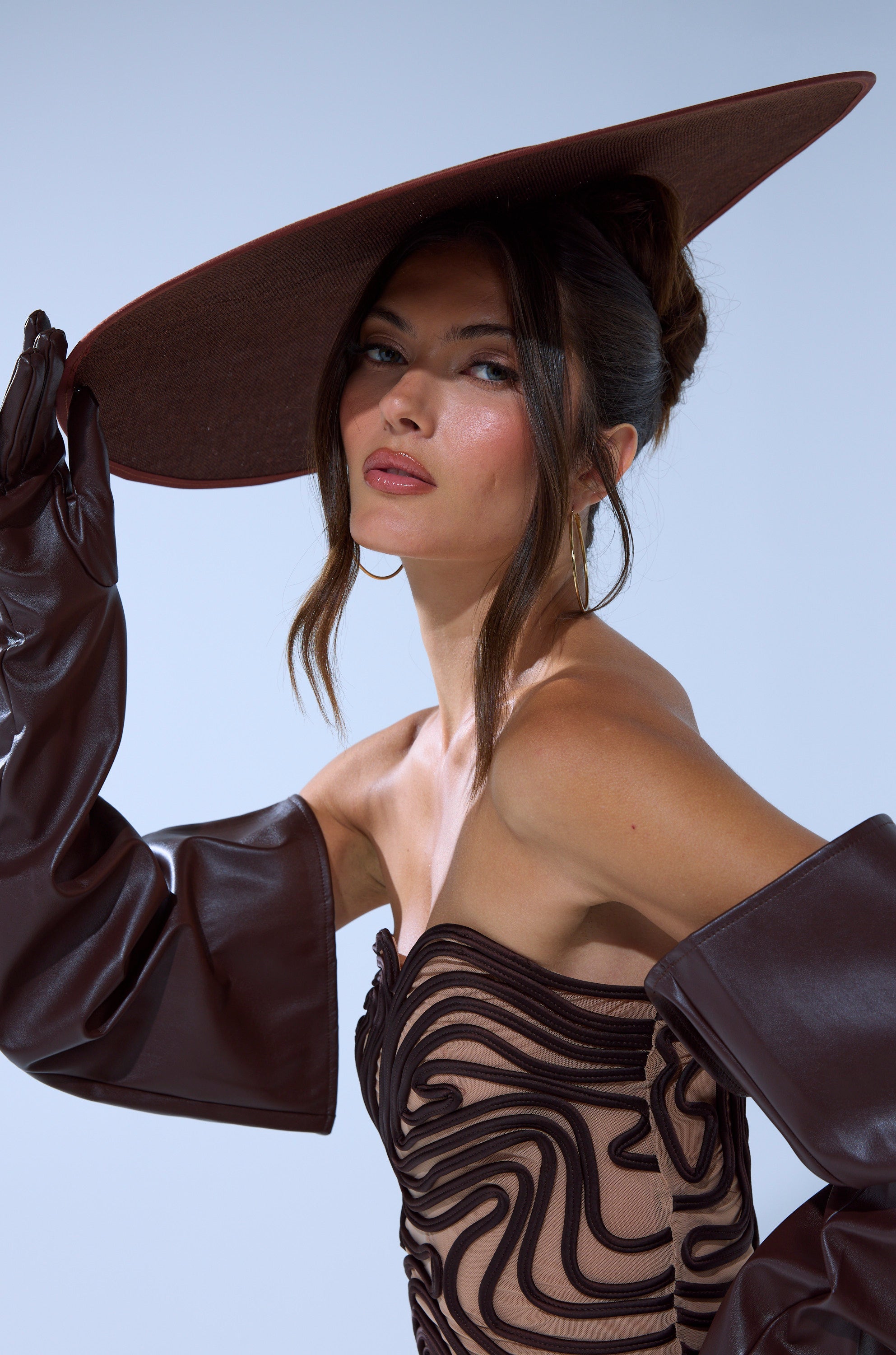 A woman poses confidently in the TOO CHIC CLIP ON HAT, paired with brown gloves and a strapless dress featuring a wavy black pattern, looking directly at the camera against a plain, light background. 2