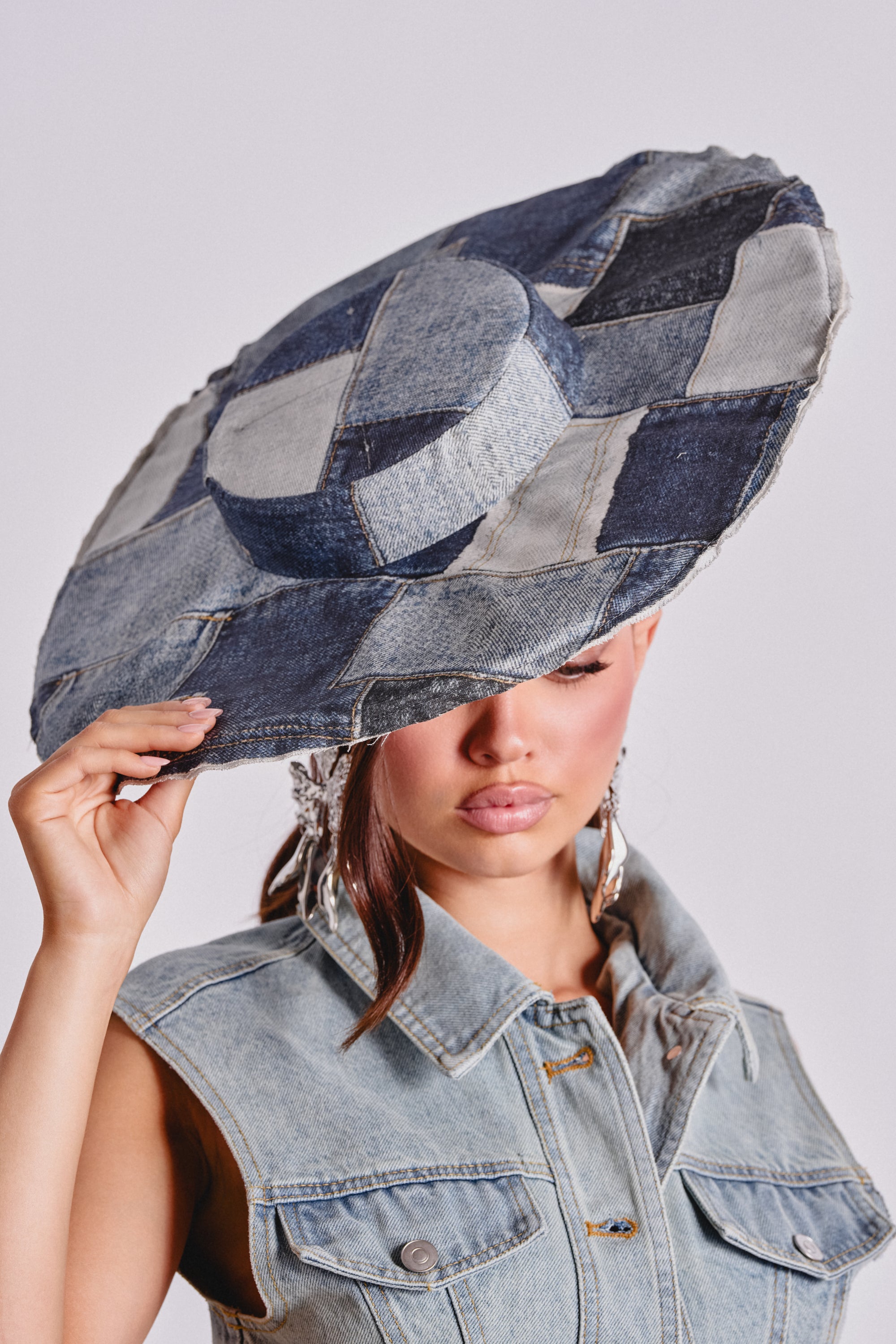 A person in a sleeveless denim jacket wears the SWEETIE DENIM BRIM HAT, holding its wide brim to partly cover their face. The plain, light background makes this bold street style accessory stand out.
