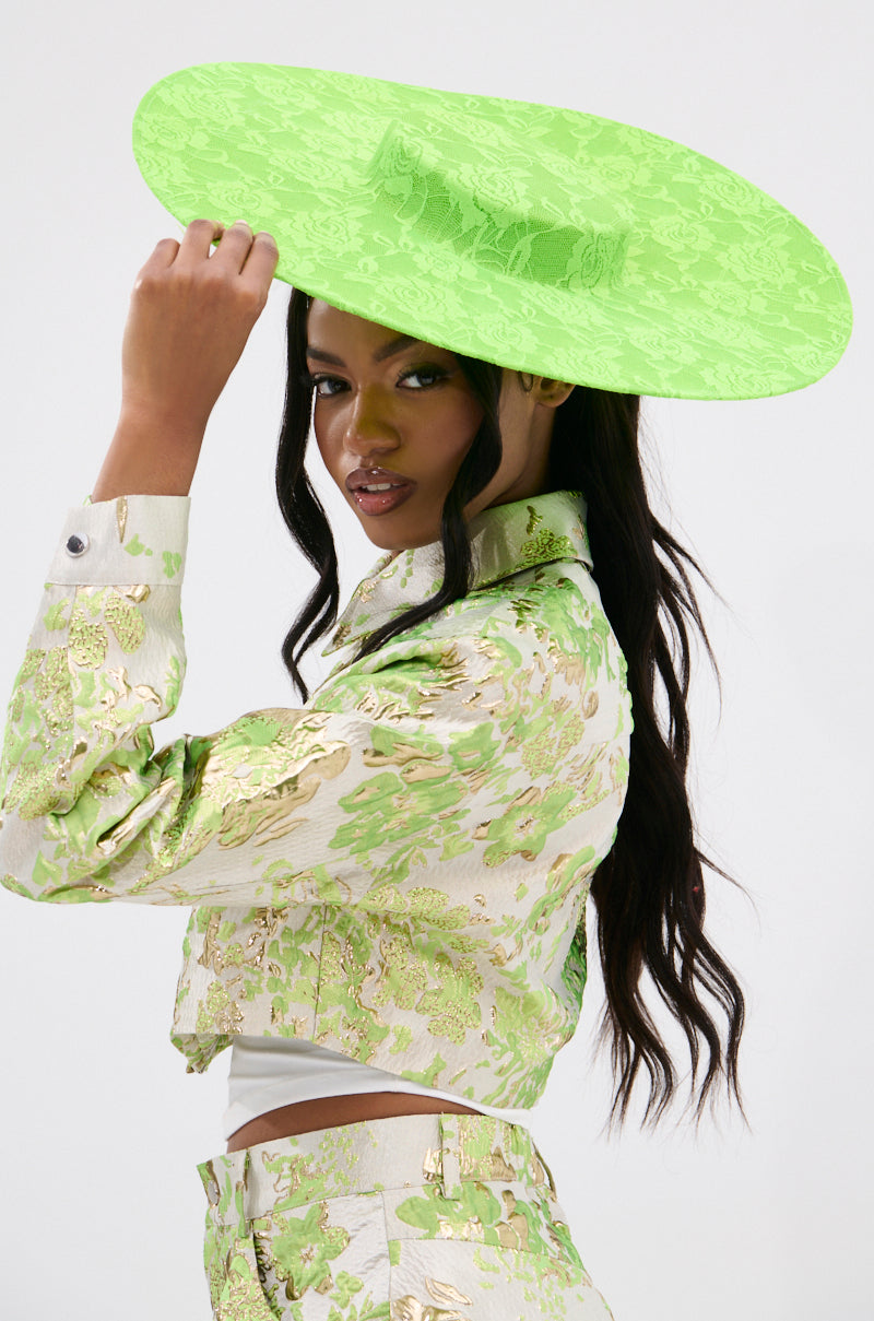 A confident woman poses, wearing the neon green "IN THE GARDEN CLIP ON HAT," perfectly complementing her floral-patterned outfit with gold accents. Her long, dark hair cascades down her back as she glances to the side.
