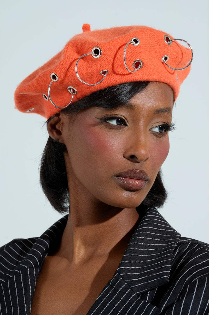 Wearing the COME ALIVE GROMMET BERET, a person pairs it flawlessly with a pinstripe blazer. Their bold blush makeup enhances the chic outfit as they look slightly to the side against a simple, light-colored backdrop.