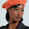 Wearing the COME ALIVE GROMMET BERET, a person pairs it flawlessly with a pinstripe blazer. Their bold blush makeup enhances the chic outfit as they look slightly to the side against a simple, light-colored backdrop.