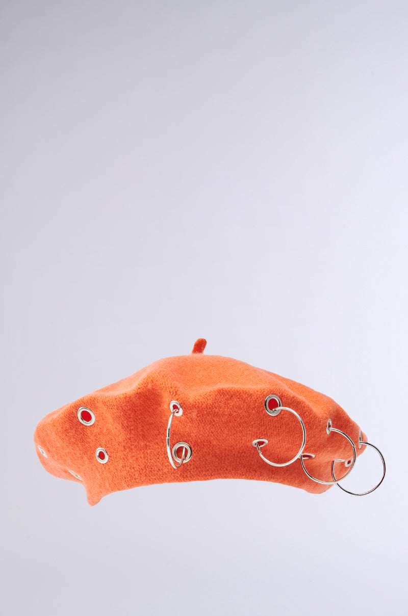 The COME ALIVE GROMMET BERET, an orange knit accessory featuring silver grommets and O-ring embellishments of varying sizes on the side, stands out stylishly against a plain gray background.