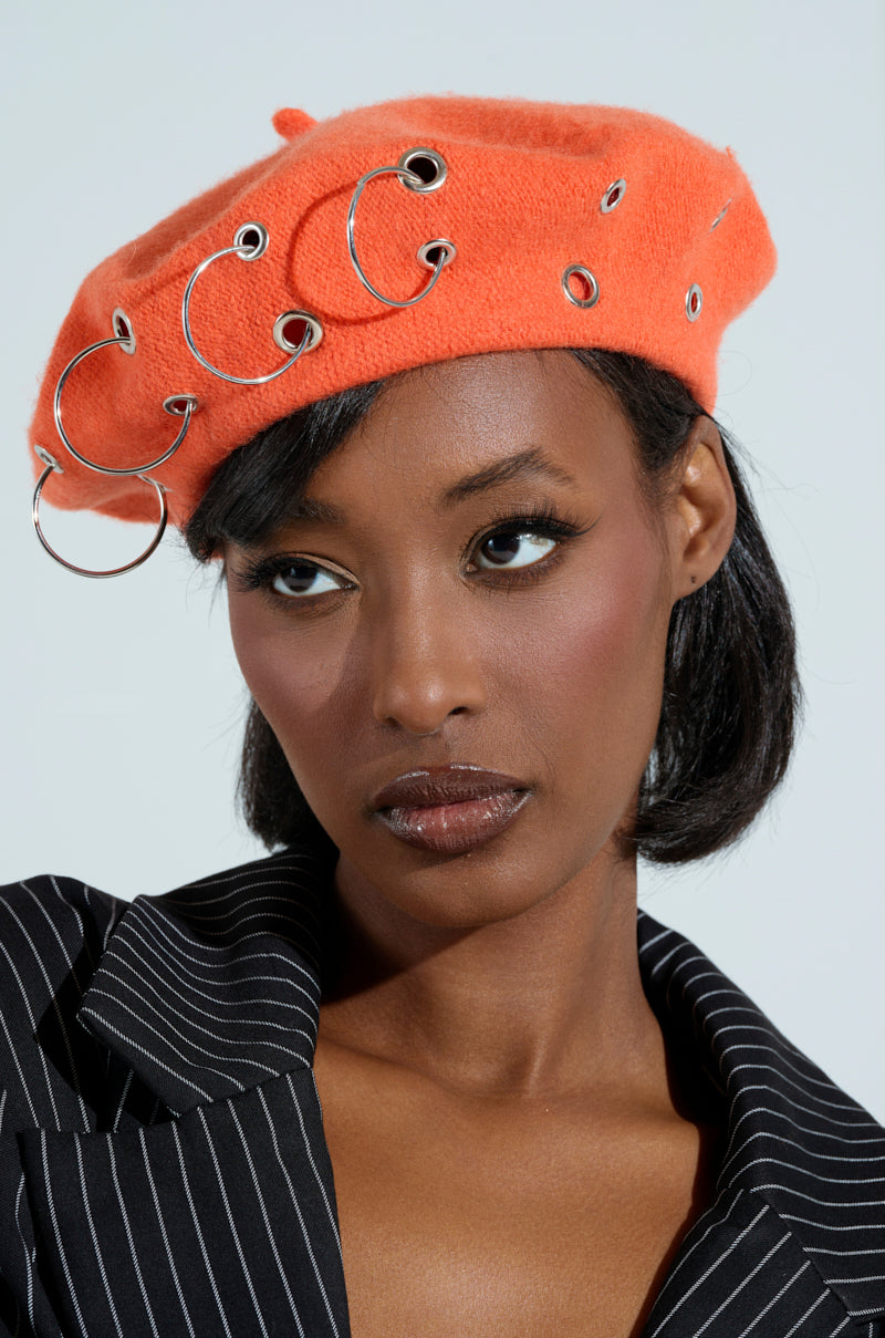 Wearing the COME ALIVE GROMMET BERET in orange, adorned with silver grommets, a person in a pinstripe suit jacket gazes seriously to the side against a plain background.