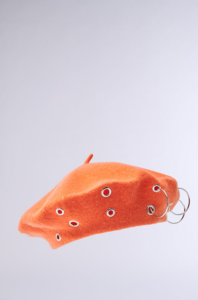 The COME ALIVE GROMMET BERET is orange and knit, adorned with multiple silver grommets and two O-ring embellishments, all shown against a plain light gray background.