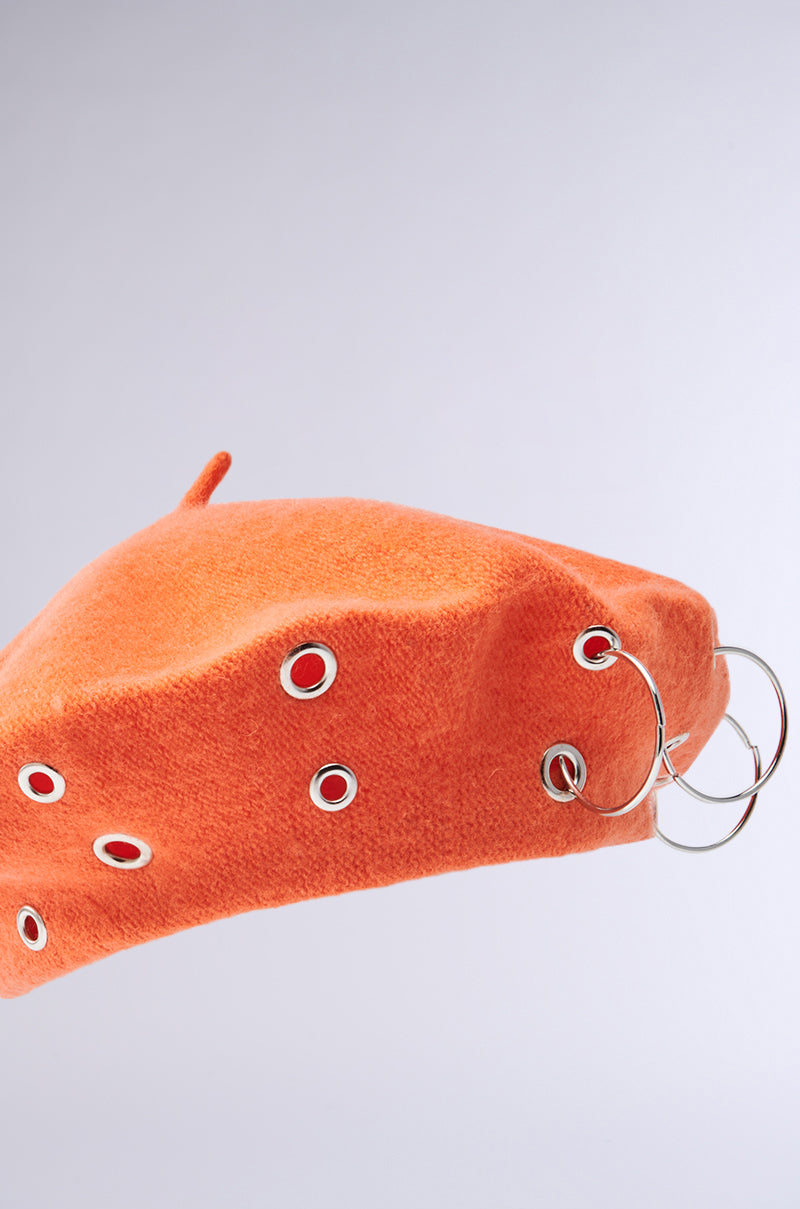 The COME ALIVE GROMMET BERET, an orange knit design, features silver grommets and two large metal rings on its side. It stands out against a light gray background, slightly tilted to the left to display its chic details.