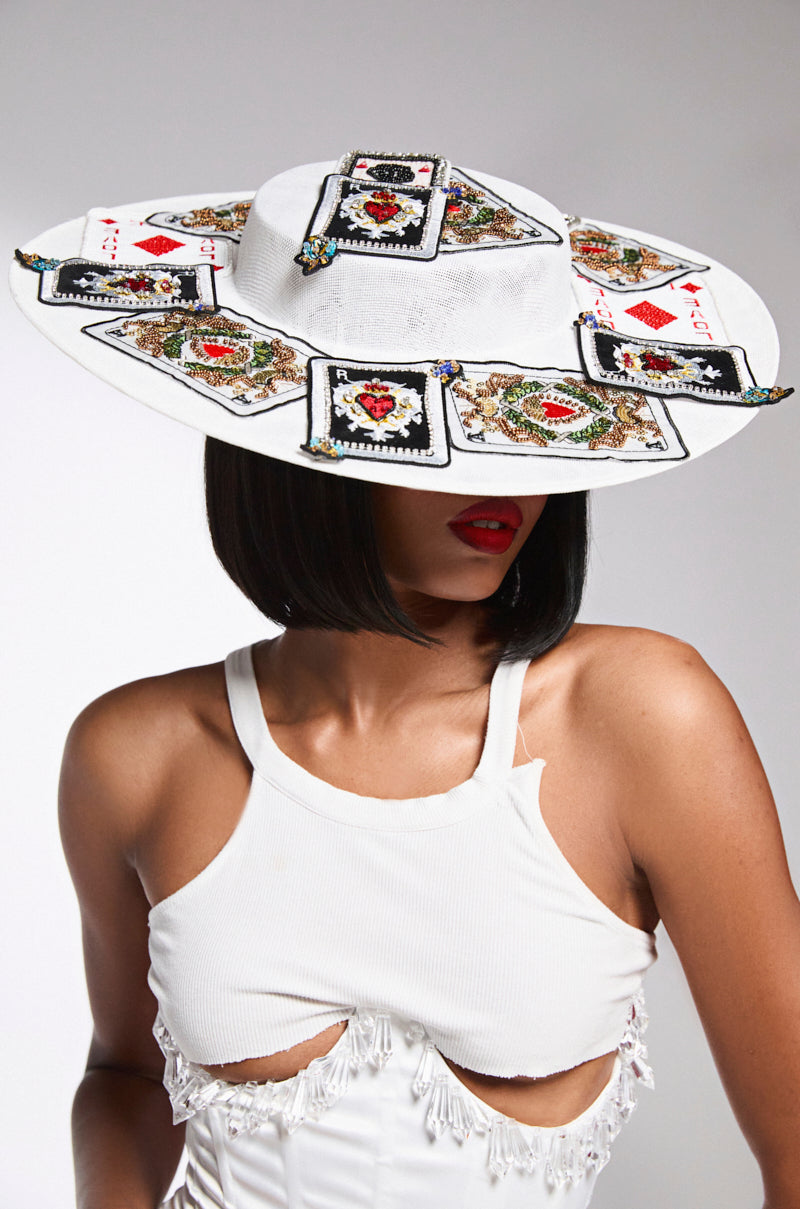 A woman with a short bob wears the POKER FACE CLIP ON HAT, a large and stylish white clip-on hat featuring playing card designs. She's in a fringed white top with red lips.