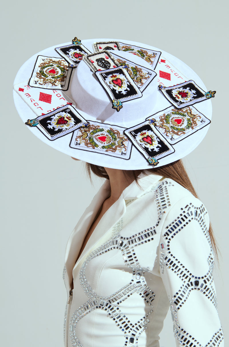 Wearing a white outfit with geometric patterns, an enigmatic figure conceals their poker face under the POKER FACE CLIP ON HAT, decorated with playing card patches. The plain, neutral background adds to the mystery.