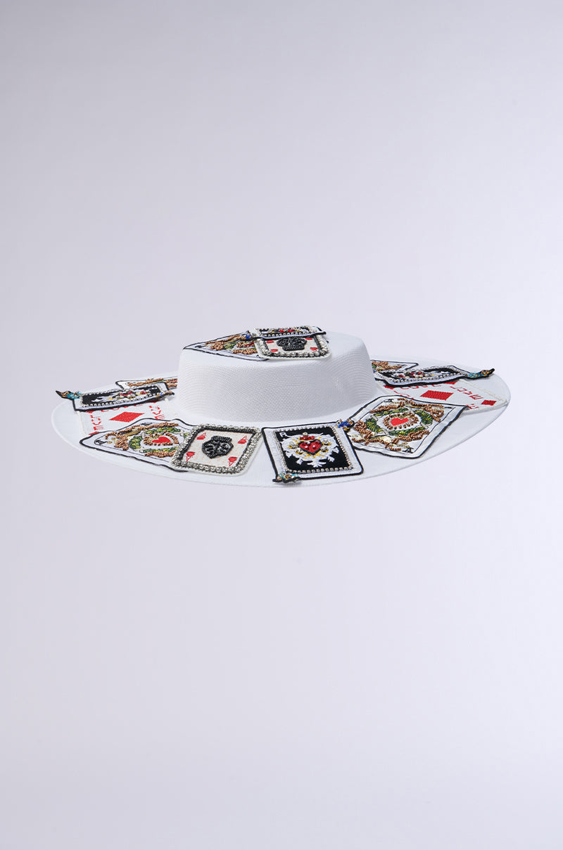 Discover the POKER FACE CLIP ON HAT: a white wide-brimmed hat featuring playing card patches with spades, hearts, diamonds, and clubs in a circular pattern around the brim, offering a unique and playful charm.