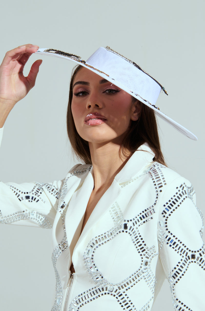 A person with long hair and glossy pink lips lifts the brim of a POKER FACE CLIP ON HAT while wearing a matching jacket with geometric mirror designs, posing against a neutral background.