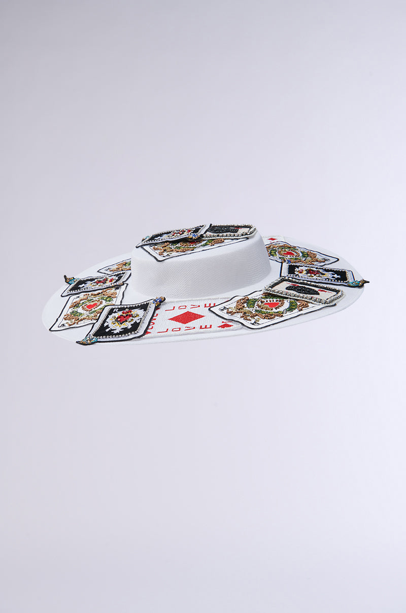 The POKER FACE CLIP ON HAT is a white wide-brimmed hat featuring colorful playing card patches and intricate embroidery on the brim. Its neutral background perfectly highlights the detailed embellishments.