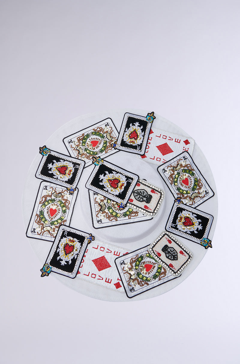 The POKER FACE CLIP ON HAT showcases a circular design with embroidered fabric resembling playing card patches. These cards feature intricate heart and floral patterns bordered in black or white, centered around layered fabric circles for a stylish and elegant look.