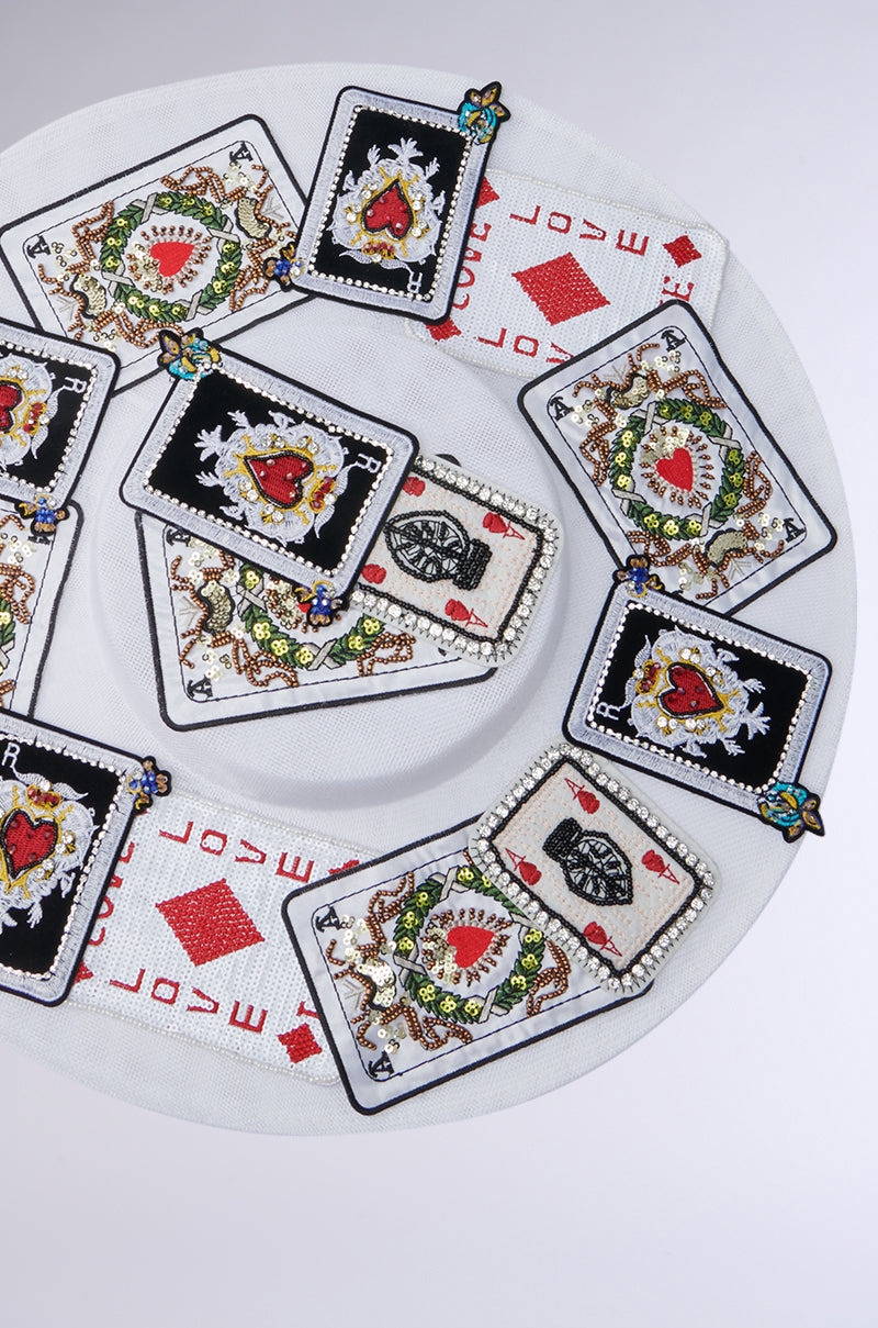A circular arrangement of intricately embroidered "POKER FACE CLIP ON HAT" patches displays hearts, flowers, and artistic designs on a round white surface with red diamonds and the word "LOVE" repeated around the edge.