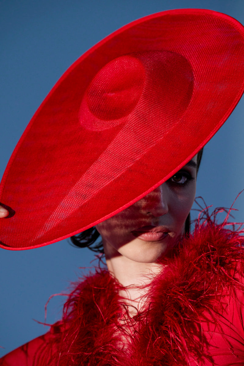 A woman wears the BLUSHING EDITORIAL CLIP ON HAT, partially covering her face and casting a shadow, paired with a bold red feathered ensemble against a vivid blue background.