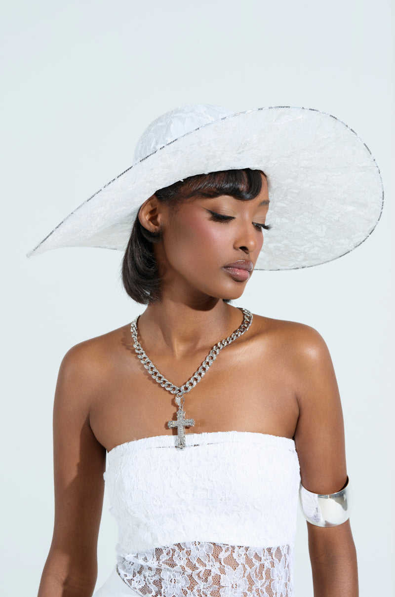 A woman poses gracefully against a light background, wearing an I AM IT LACE HAT, a strapless floral lace top, a large silver chain necklace with a cross pendant, and a silver arm cuff. Her eyes are closed.