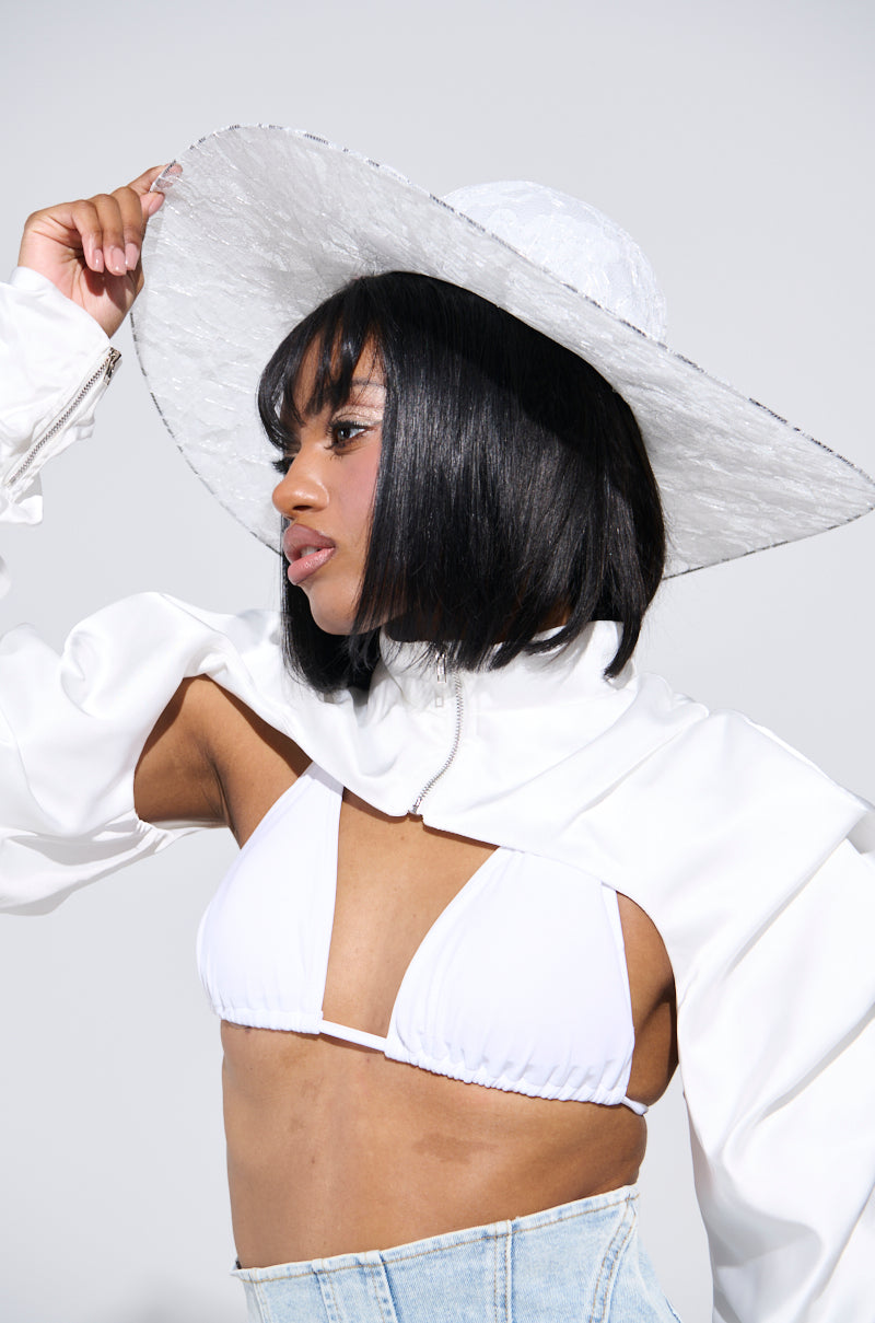 Wearing a wide-brim white I AM IT LACE HAT, a person with a bob hairstyle dons a stylish white outfit featuring a cropped top and denim piece, highlighted by dramatic sleeves. They pose elegantly against a plain background. 0