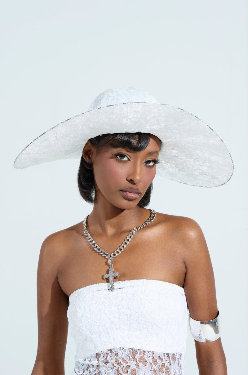 A woman in a white floral lace strapless top and an I AM IT LACE HAT poses against a light background. She wears a silver chain necklace with a cross and a silver armband, exuding calm and confidence.