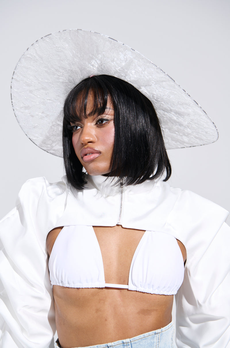 A person with straight black hair wears the I AM IT LACE HAT—a wide-brimmed white hat with floral lace—paired with a white jacket featuring puffed sleeves and a white bikini top. They have a neutral expression against a plain background.
