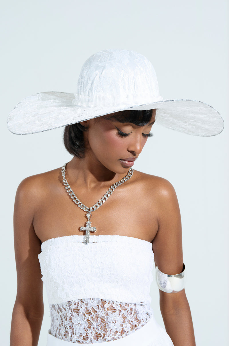 A woman in a white floral lace strapless dress wears an I AM IT LACE HAT along with a silver chain necklace featuring a cross and a silver armband. Her eyes are downcast, makeup subtle, set against a light backdrop.