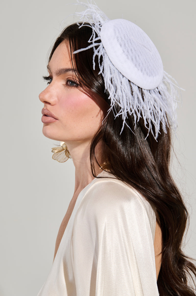 A woman with long dark hair sports a FEATHER FAN CLIP ON HAT, featuring feather trim, paired with large flower-shaped earrings. She is dressed in a light, silky top and is depicted in profile against a neutral background.
