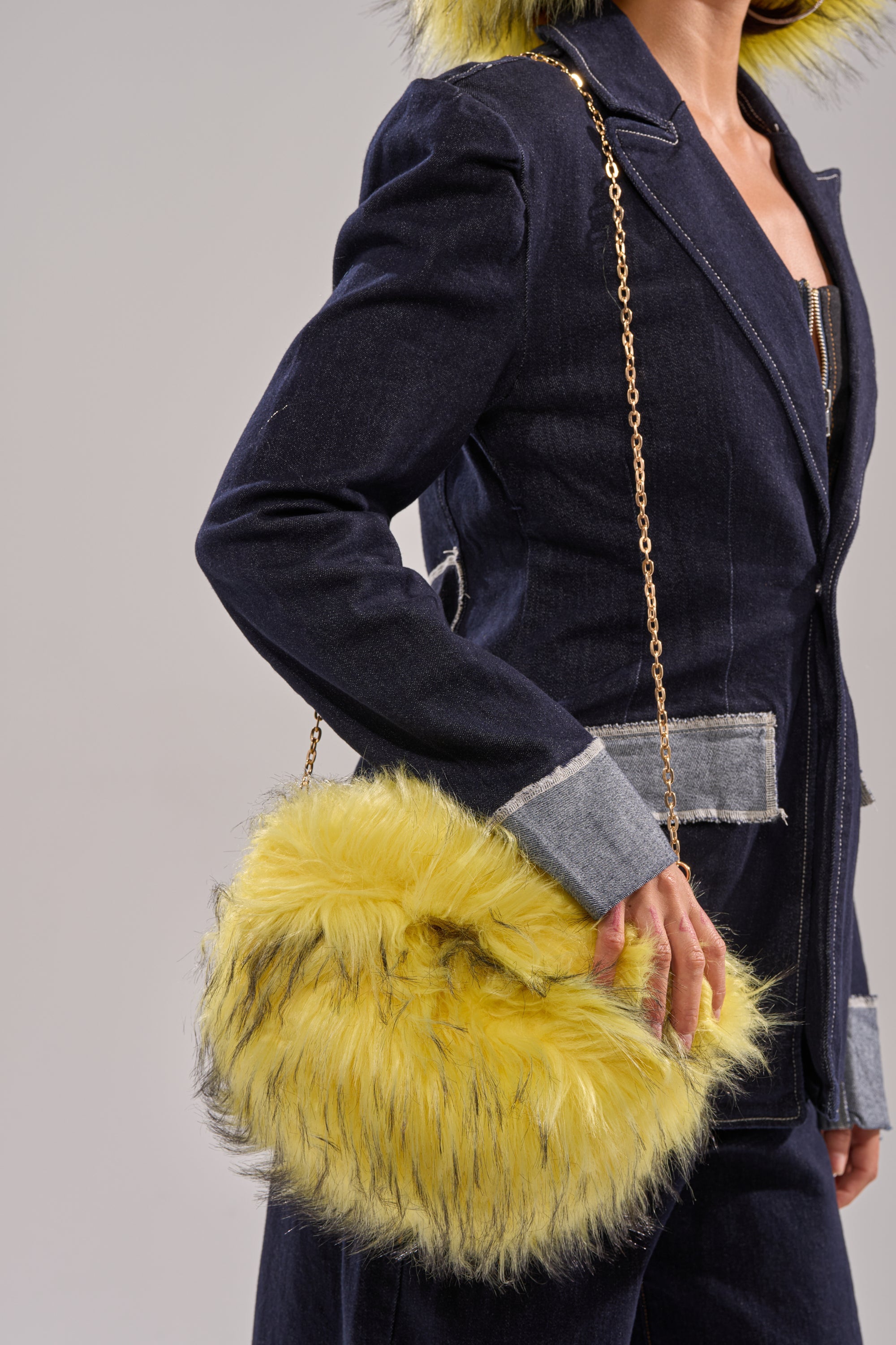 A person in a dark denim jacket with gray accents holds the ELLIE FAUX FUR PURSE, a round, fluffy yellow bag with a gold chain crossbody strap. The person's head is partially out of frame. 1