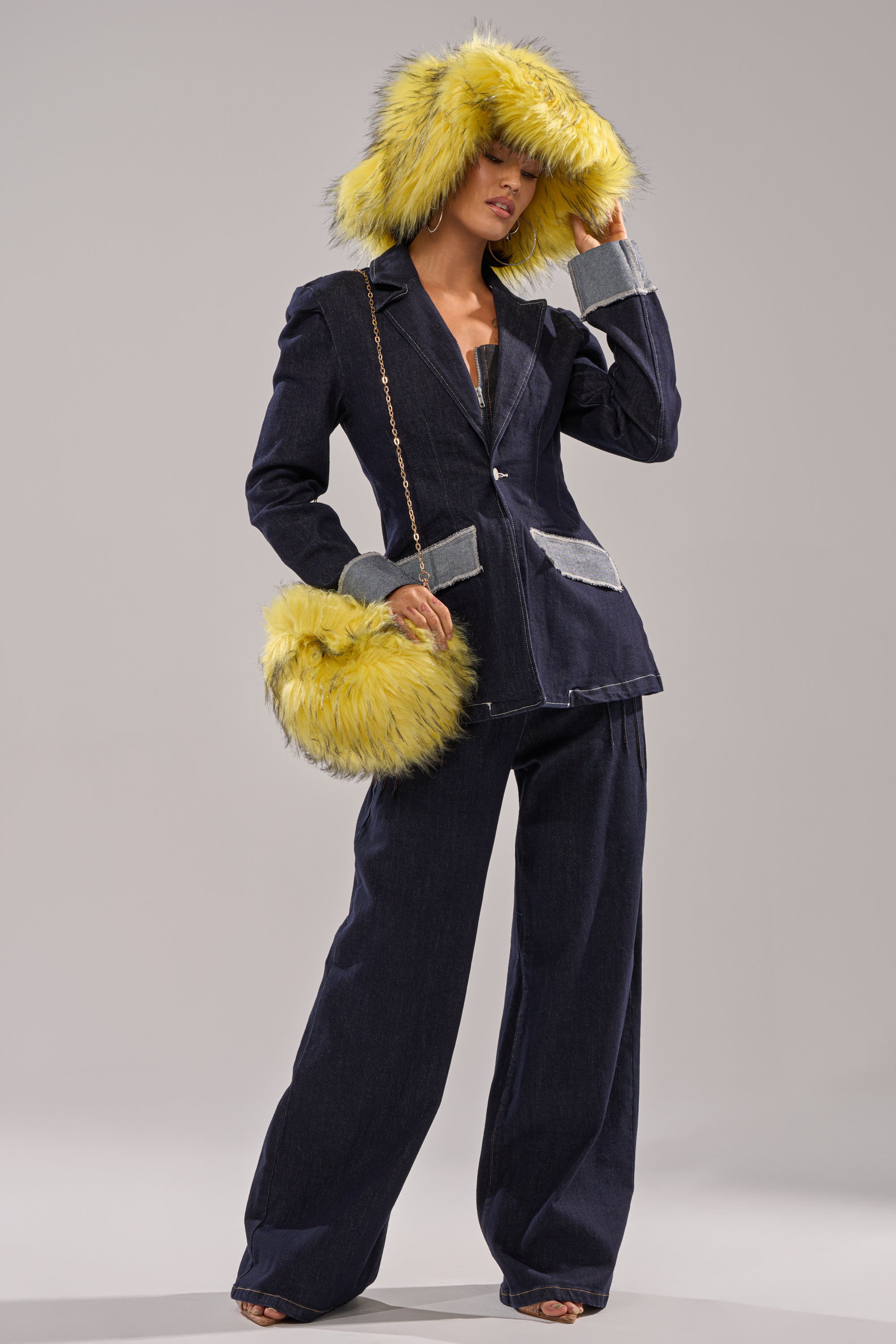 A woman poses against a plain background in a dark denim suit with wide-leg pants and a jacket featuring large gray pockets. She accessorizes with the ELLIE FAUX FUR PURSE in yellow and gray, plus a matching hat.