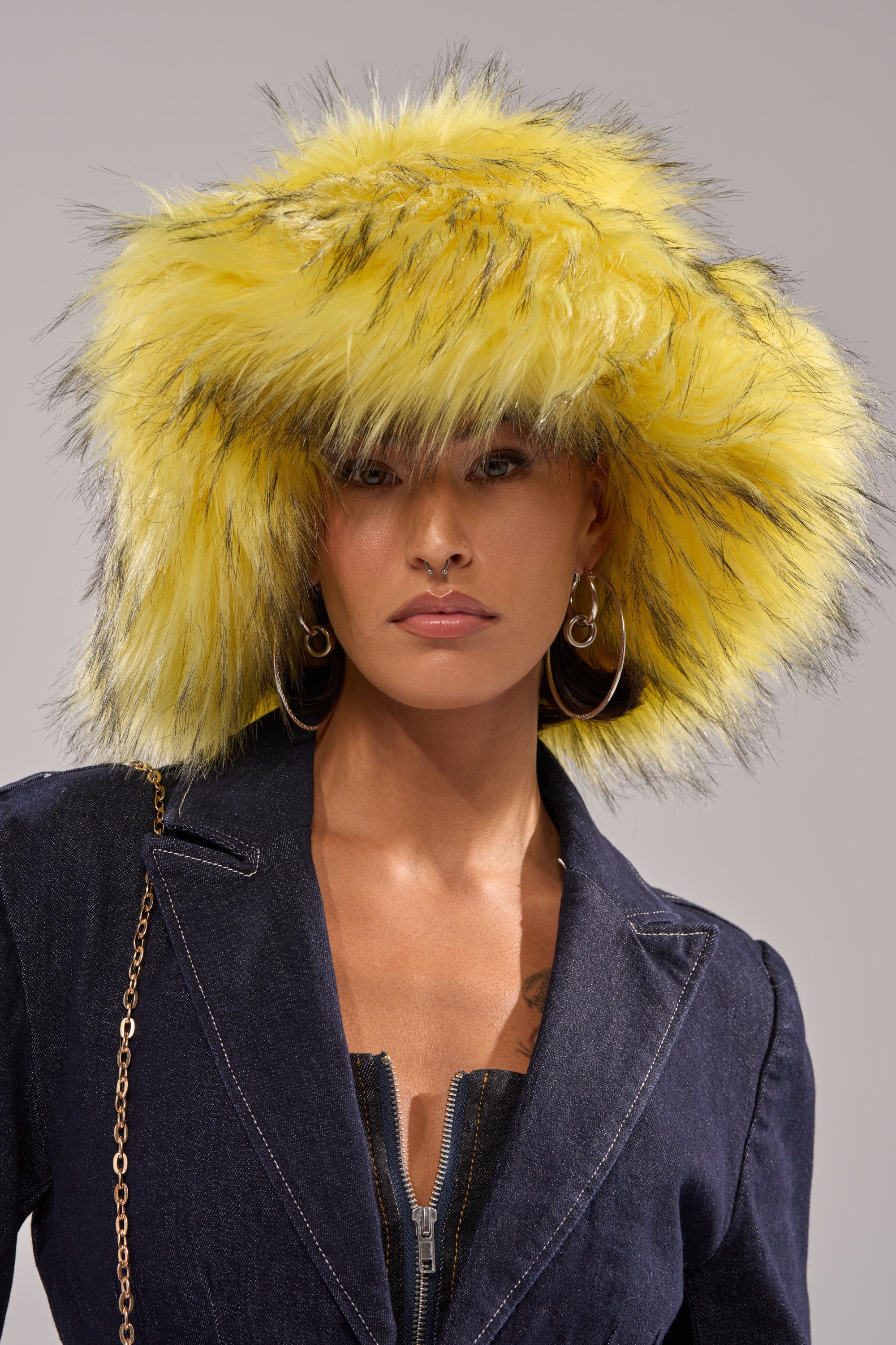 A woman wears the AKIRA Label ELLIE FAUX FUR BUCKET HAT in yellow, paired with gold hoop earrings and a dark denim jacket with zipper and chain strap, looking confidently at the camera against a neutral background. 0