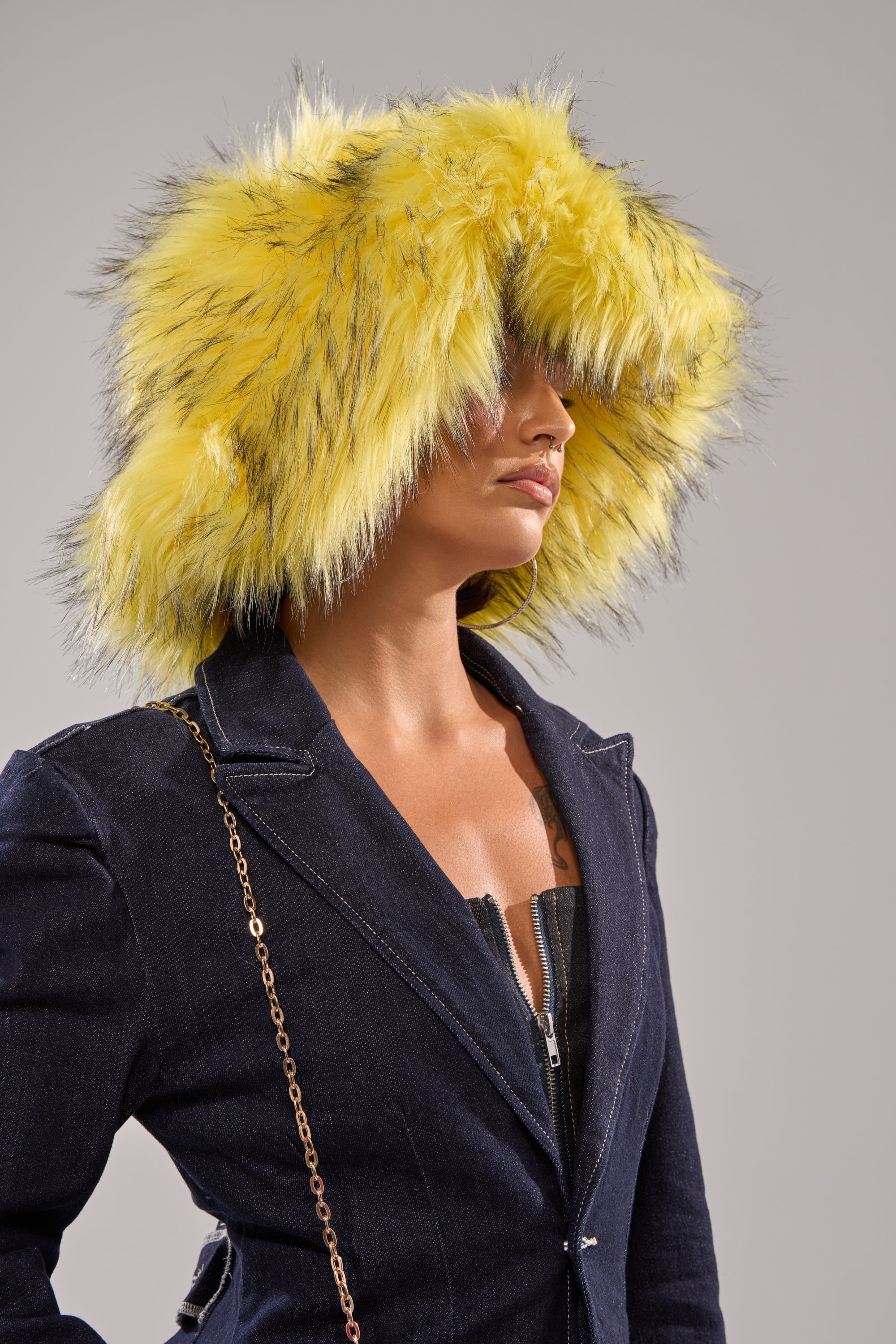 Wearing the bold ELLIE FAUX FUR BUCKET HAT in large yellow faux fur with black streaks, this person pairs it with a dark denim jacket and gold chain strap, standing against a neutral background. 1