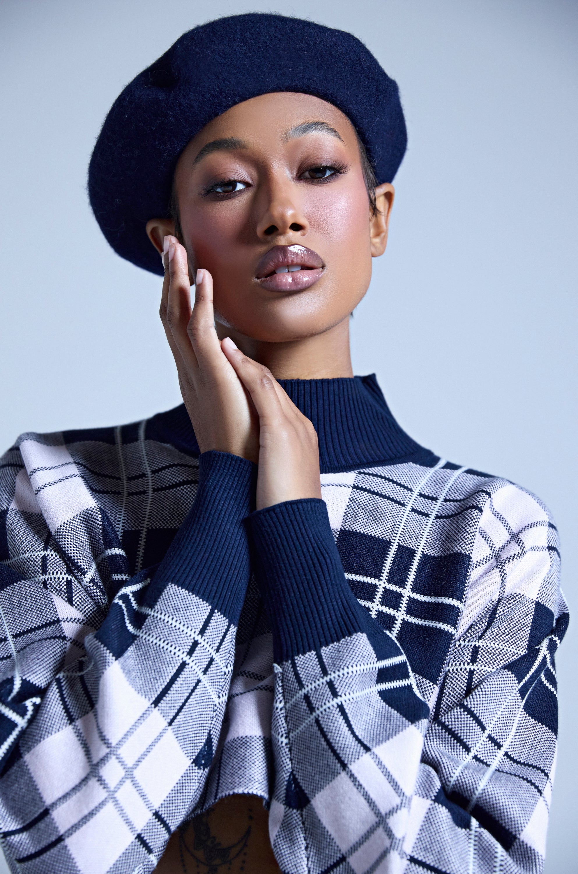 A person poses confidently against a plain light background, wearing the TOO COZY NAVY BERET and a checkered sweater, gently touching their face. The beret adds a stylish touch to the look. 1