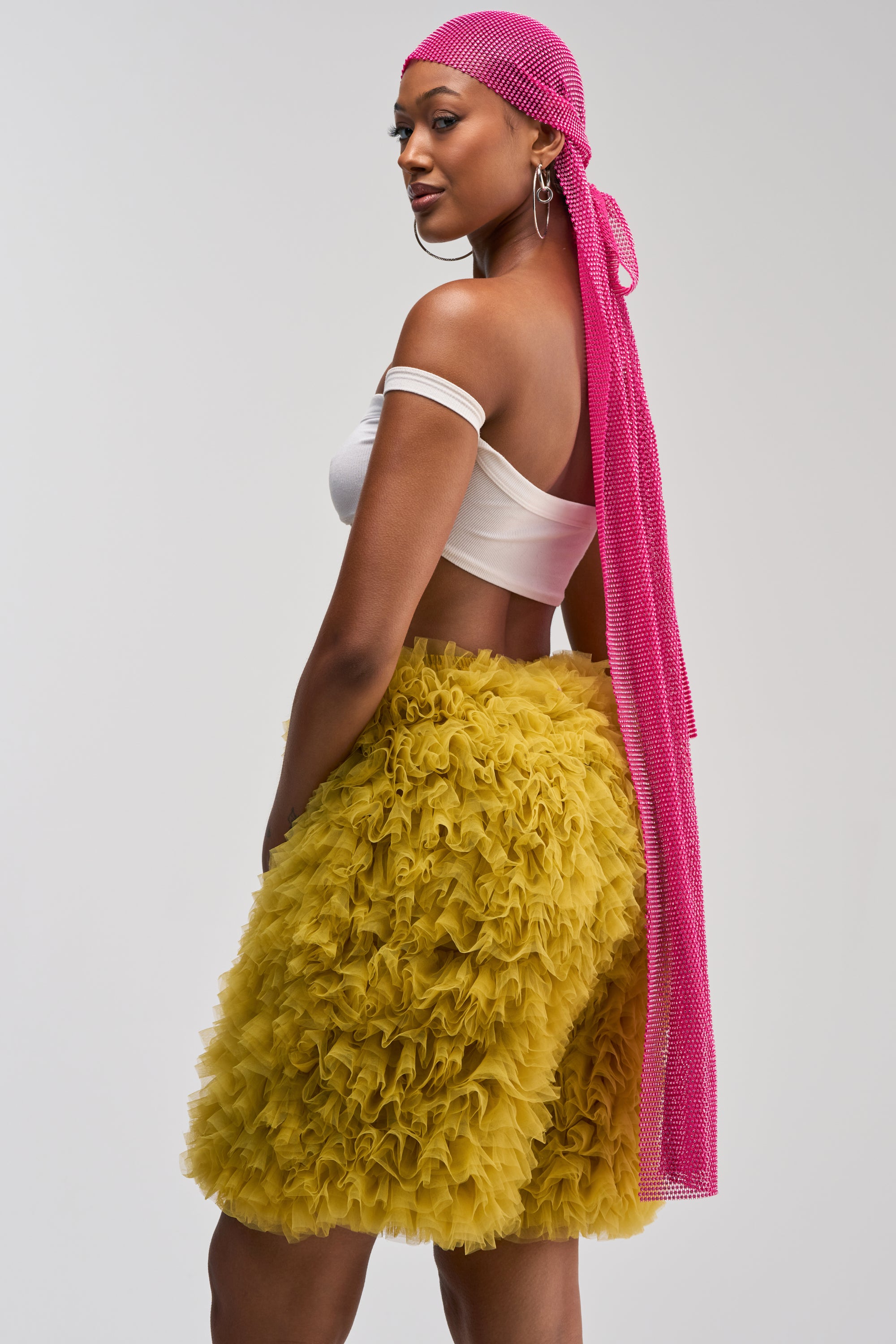 Wearing the ALL NIGHT LONG RHINESTONE DURAG, a woman in a white off-shoulder top and yellow ruffled skirt stands sideways, looking over her shoulder against a plain background, making this sparkling festival durag the perfect statement summer accessory.