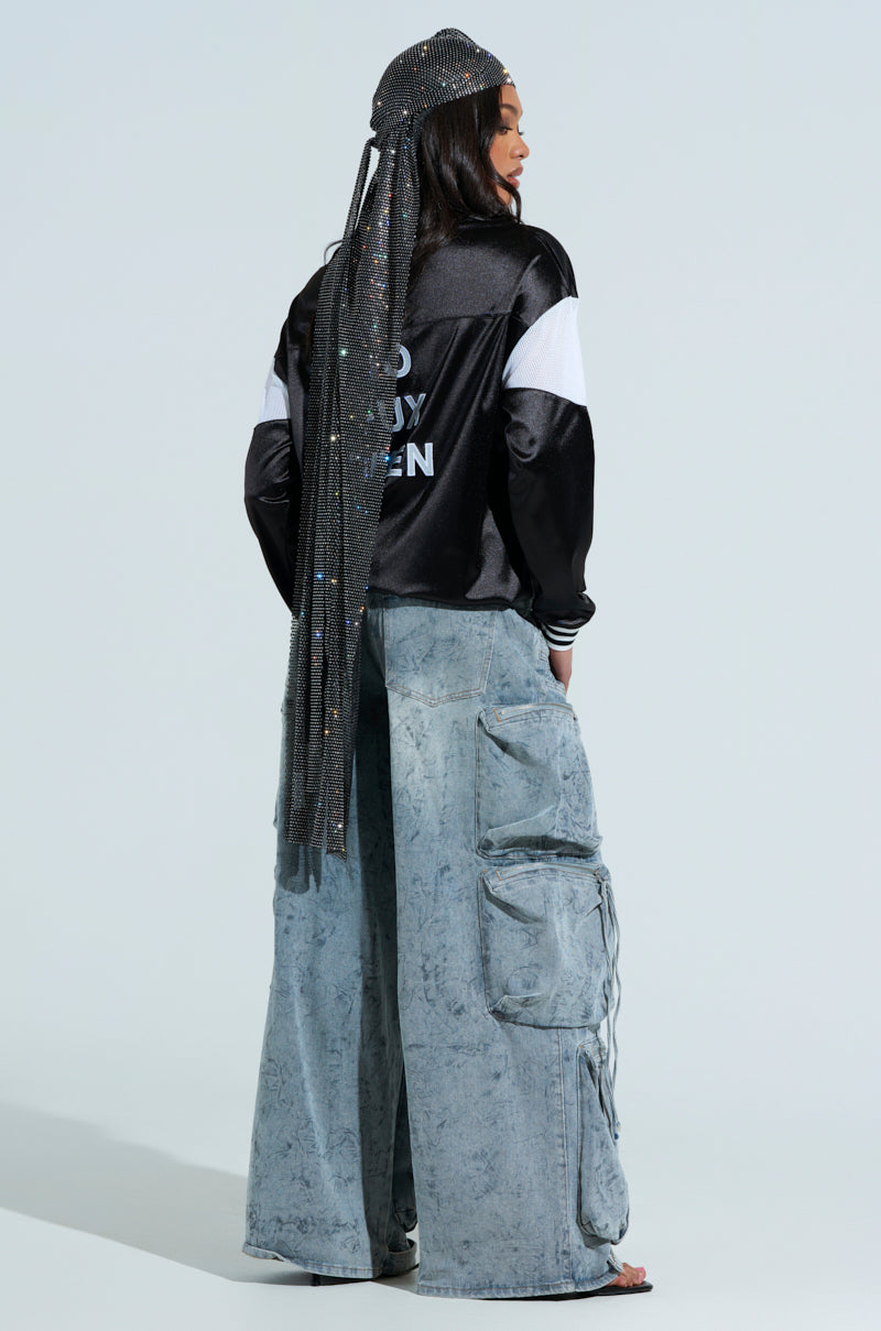 Their back to the camera, they wear a black jacket with white text and oversized light blue cargo pants. Their long, sparkling headscarf cascades down their back like the ONLY THE ELITE RHINESTONE NET DURAG against a plain, light-colored background.
