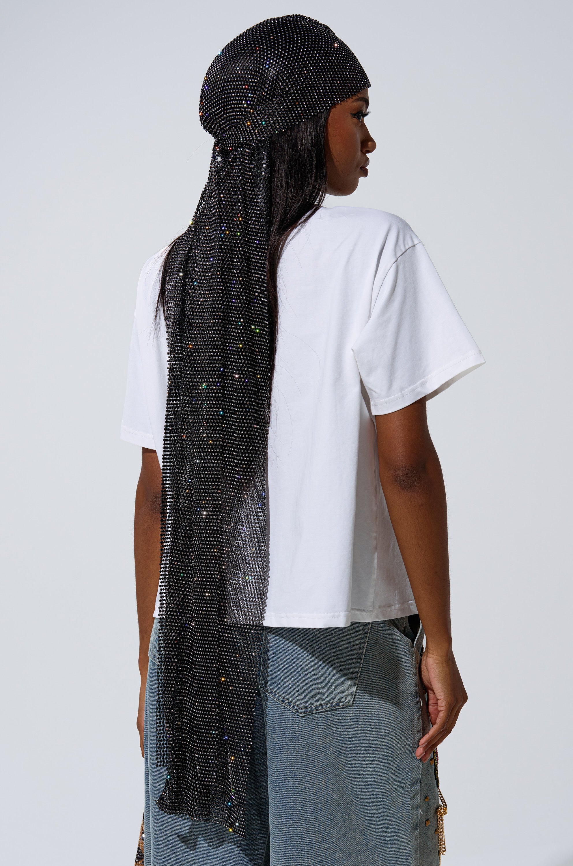 A person stands with their back to the camera, wearing a white t-shirt, light blue jeans, and the ONLY THE ELITE RHINESTONE NET DURAG adorned with sparkling jewels elegantly draping down their back.
