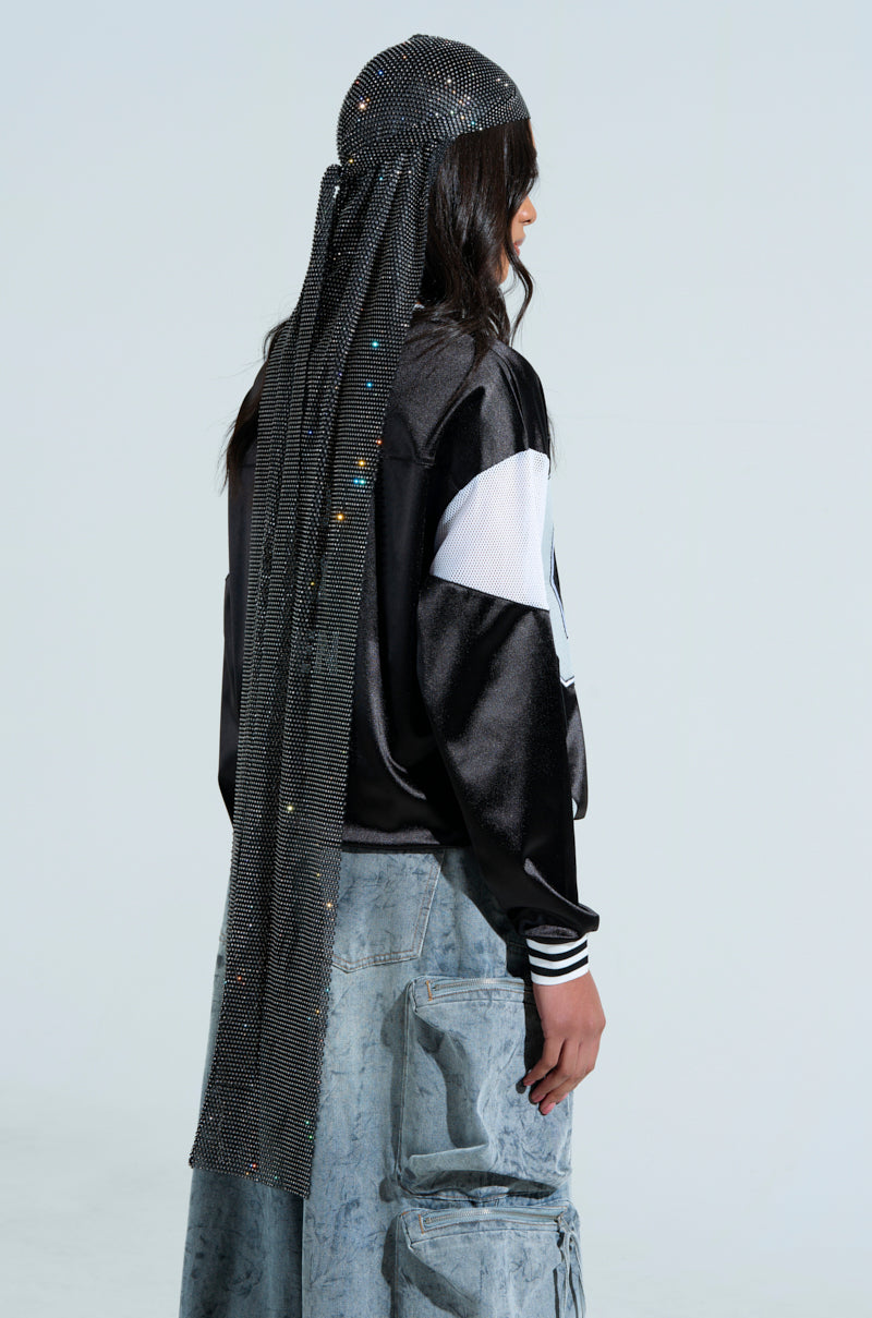 A person with long hair wears the ONLY THE ELITE RHINESTONE NET Durag and a black and white jacket. They sport light blue jeans with large pockets, standing gracefully against a plain background.