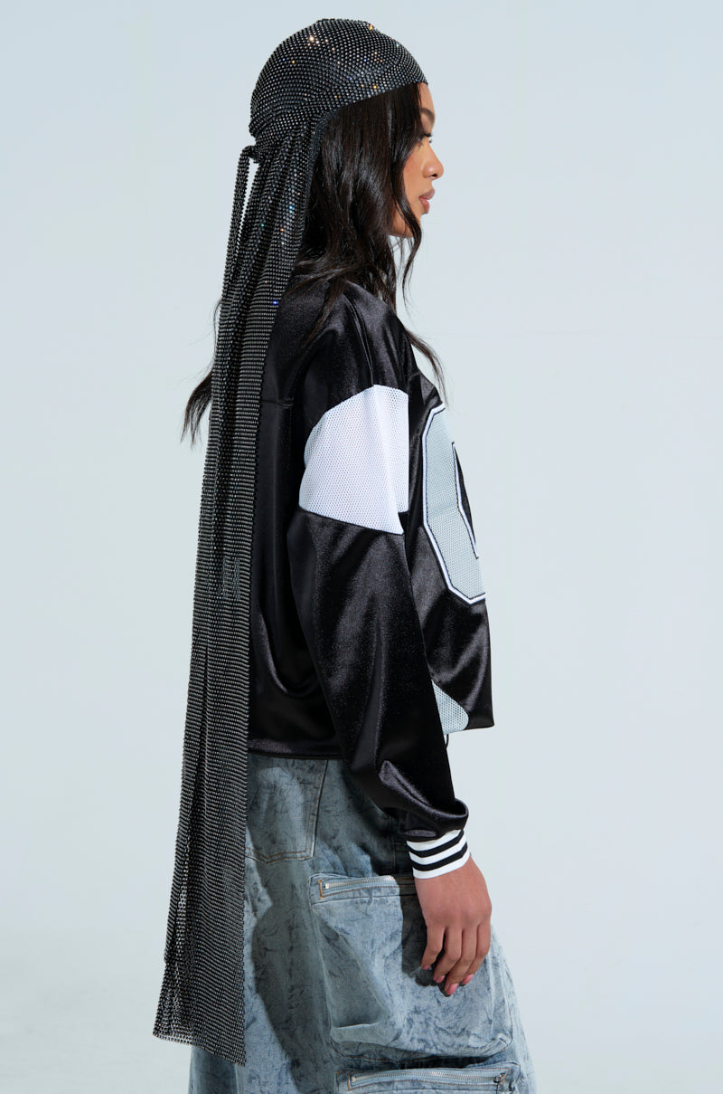 Against a neutral background, a person in a black and white sports jacket and casual blue jeans stands sideways. Their long hair is covered by the glittering ONLY THE ELITE RHINESTONE NET DURAG, resembling an embellished headscarf.