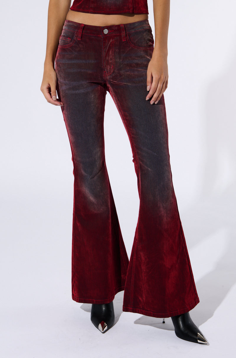 A person models the CRYSTAL VELVET DENIM PANT IN RED, characterized by its velvety fabric and bell bottom design. The high-waist pants feature front pockets and a button closure. Complementing the pants, the model wears black pointed-toe boots against a plain white background.