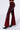 A person is modeling the CRYSTAL VELVET DENIM PANT IN RED, featuring a high-rise design with a slightly worn texture. The model pairs these red bell bottom pants with black pointed-toe shoes that have silver stiletto heels. The background is plain and light-colored.