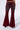 A person is wearing the CRYSTAL VELVET DENIM PANT IN RED, featuring visible seams and back pockets. They are standing against a plain white background with their back facing the camera. The wide flares of these red velvet pants give them a slightly worn look.