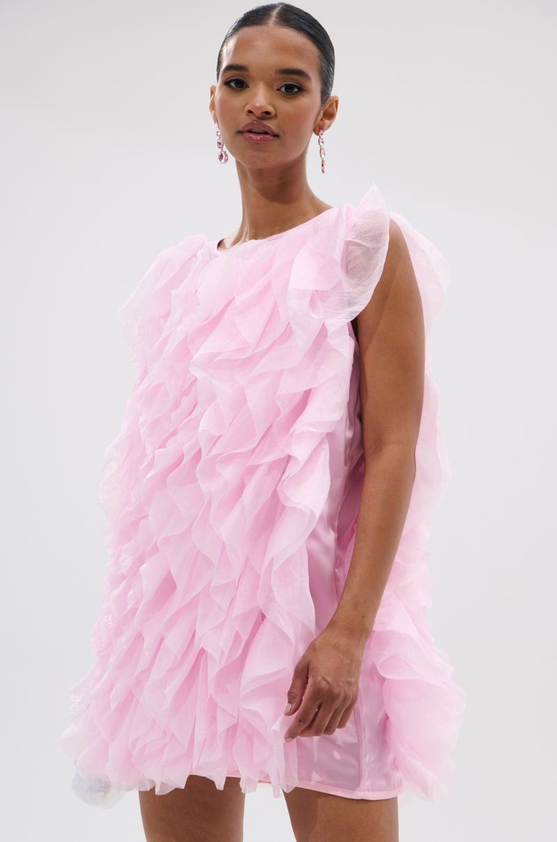 A woman stands against a plain light background, wearing the LOVER GIRL RUFFLE MINI DRESS—a sleeveless pink organza mini with layered ruffles—and matching pink drop earrings. Her hair is pulled back for an elegant look. 0