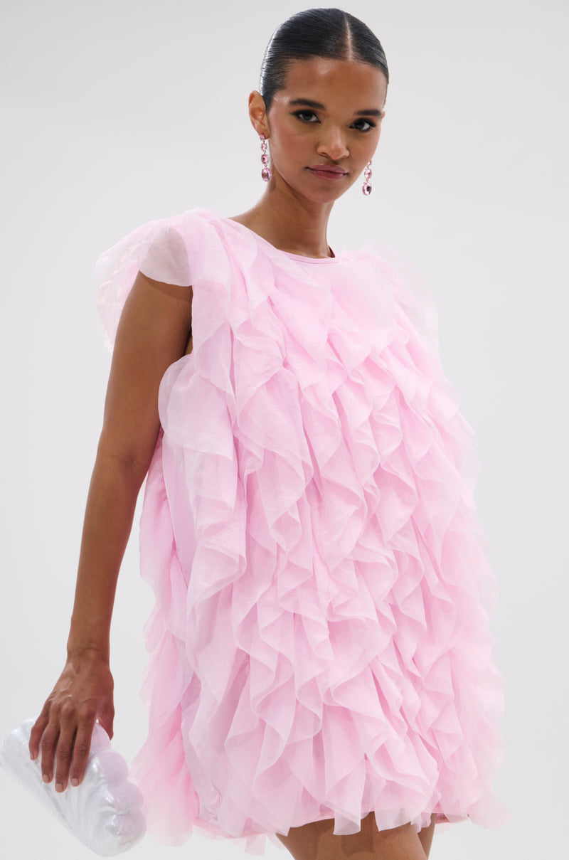 A woman wears the LOVER GIRL RUFFLE MINI DRESS in pink with a small clear clutch, drop earrings, and slicked-back hair against a plain light background. 1