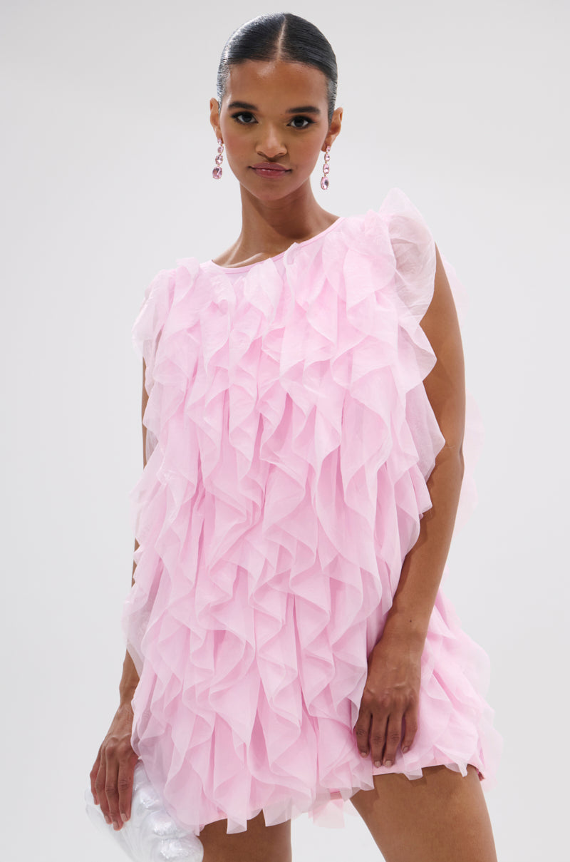 A woman stands against a plain light background, wearing the LOVER GIRL RUFFLE MINI DRESS with pink drop earrings and holding a white clutch bag. 2