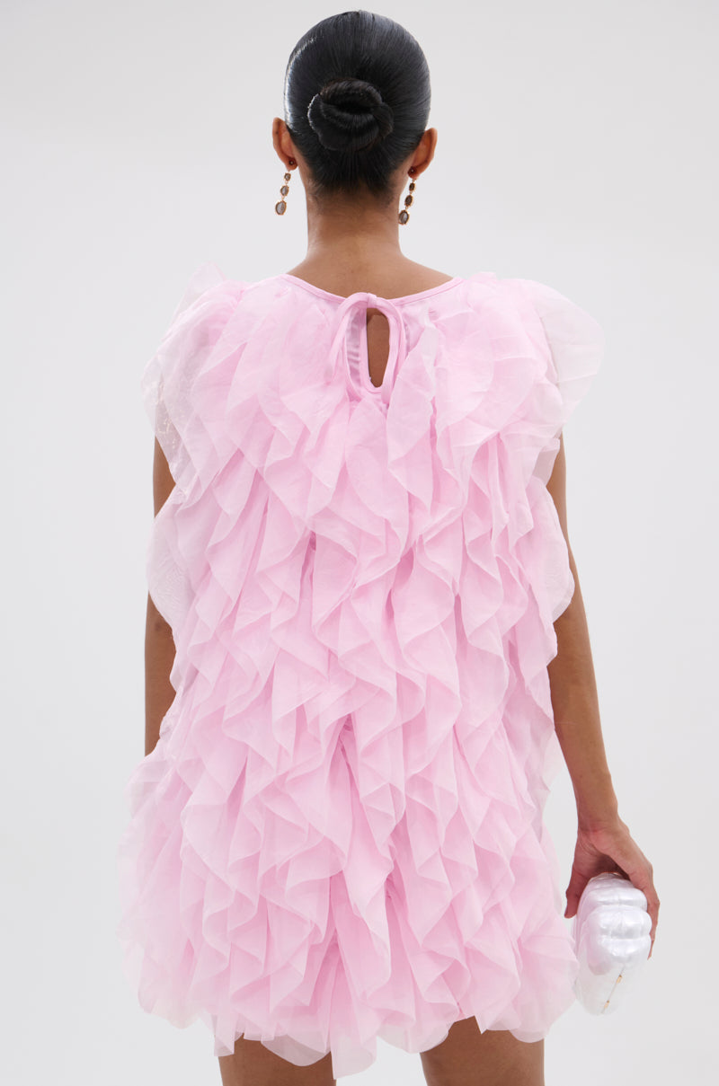 A woman with a sleek bun seen from behind wears the LOVER GIRL RUFFLE MINI DRESS in pink organza with layered ruffles, holding a small white clutch in her right hand against a plain light background. 4