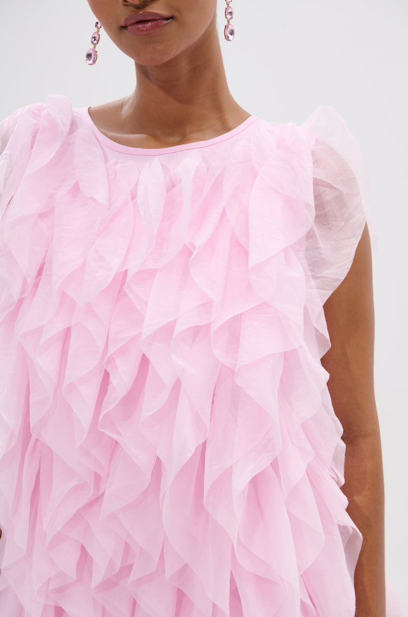 A woman wears the LOVER GIRL RUFFLE MINI DRESS, a sleeveless, sheer light pink organza mini with ruffles and statement earrings, pictured from shoulders to mid-torso against a plain background. 7