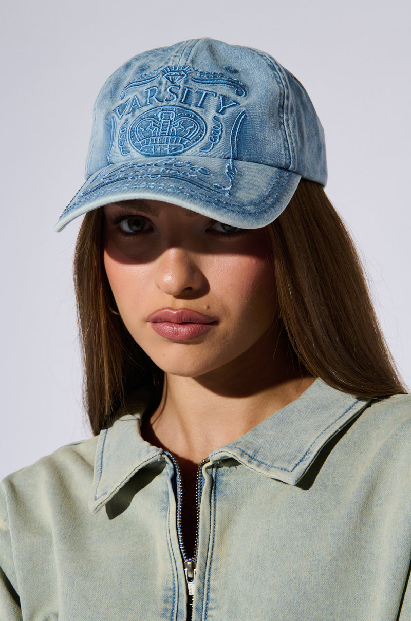 A person with long brown hair wearing the MILAN LOVE SNAPBACK HAT, featuring an acid wash denim design and the word "Varsity" embroidered on it. They are also wearing a light blue denim jacket and looking directly at the camera. The background is plain and light-colored.