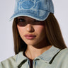 A person with long brown hair wearing the MILAN LOVE SNAPBACK HAT, featuring an acid wash denim design and the word "Varsity" embroidered on it. They are also wearing a light blue denim jacket and looking directly at the camera. The background is plain and light-colored.