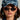 Wearing the DENIM SNAPBACK with a textured finish and adjustable buckle strap, along with dark oval sunglasses, large hoop earrings, and a white jacket, the person with long dark hair gazes directly at the camera against a plain background.